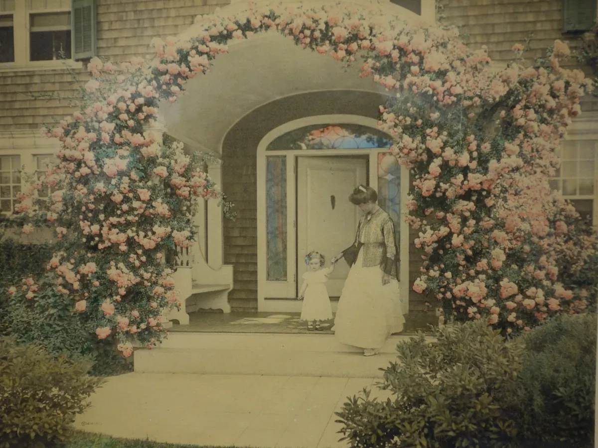 A hand-colored early 1900s photograph of a mother and child under a rose-covered arch