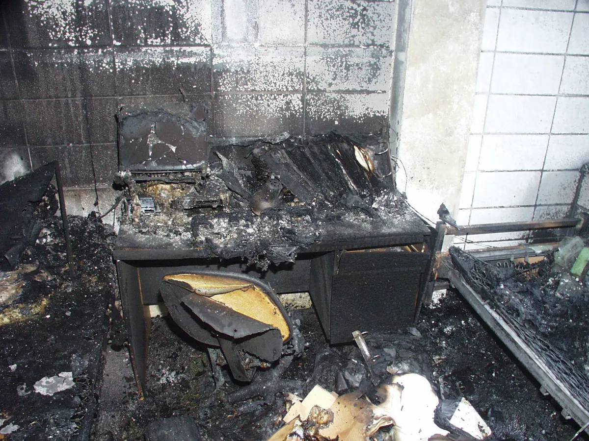 A fire-damaged room with smoke-blackened walls and a charred desk and chair